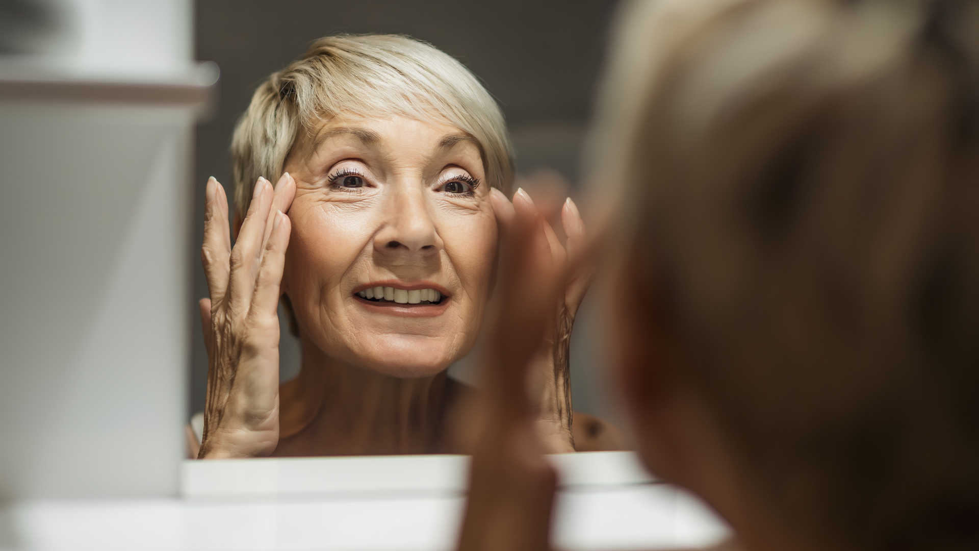 an older woman looks into a mirror with her hands grabbing at the loose skin around her eyes and face
