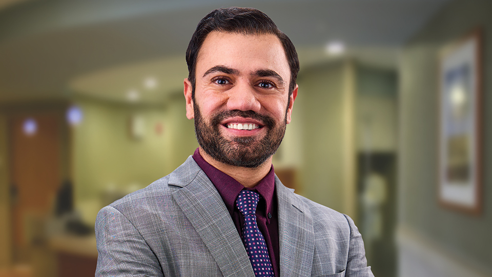 Dr. Salman Nusrat smiles in a tie and blazer with a clinical background pictured behind him