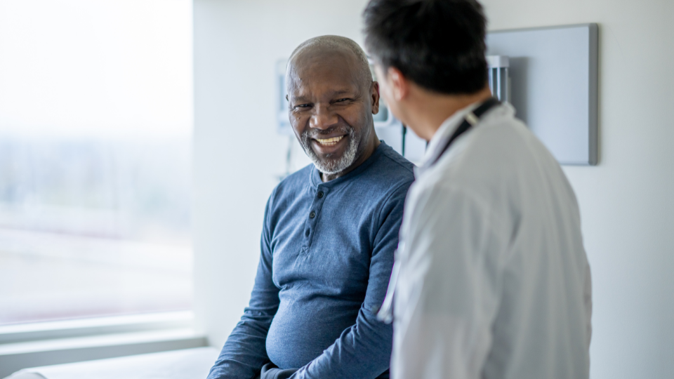 A senior gentleman of African decent sits up on an exam table during a routine medical exam. His doctor is standing to the side of him in a white lab coat as they discuss his health.