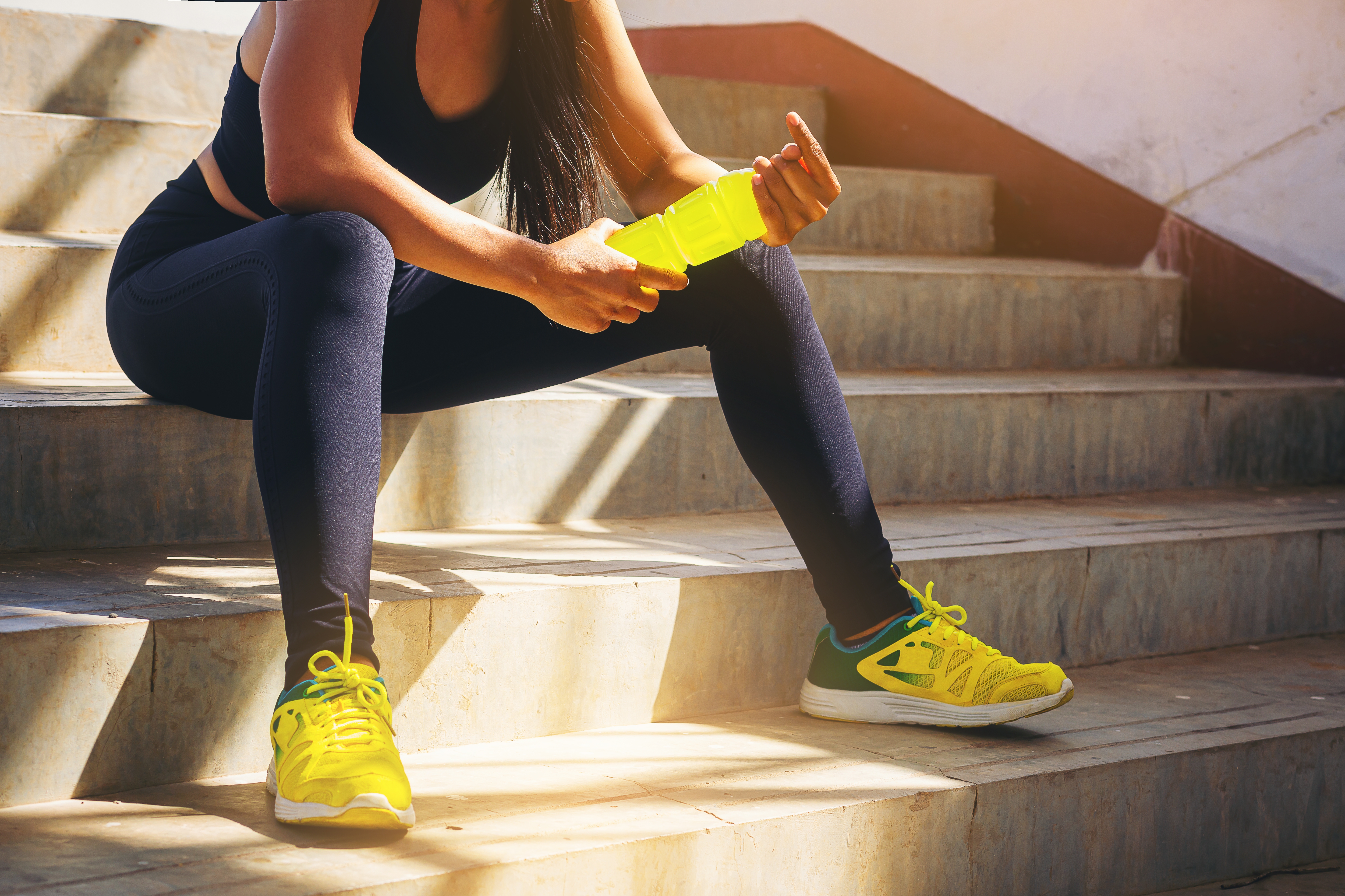 A woman sits on the steps holding a bottle filled with a yellow electrolyte drink