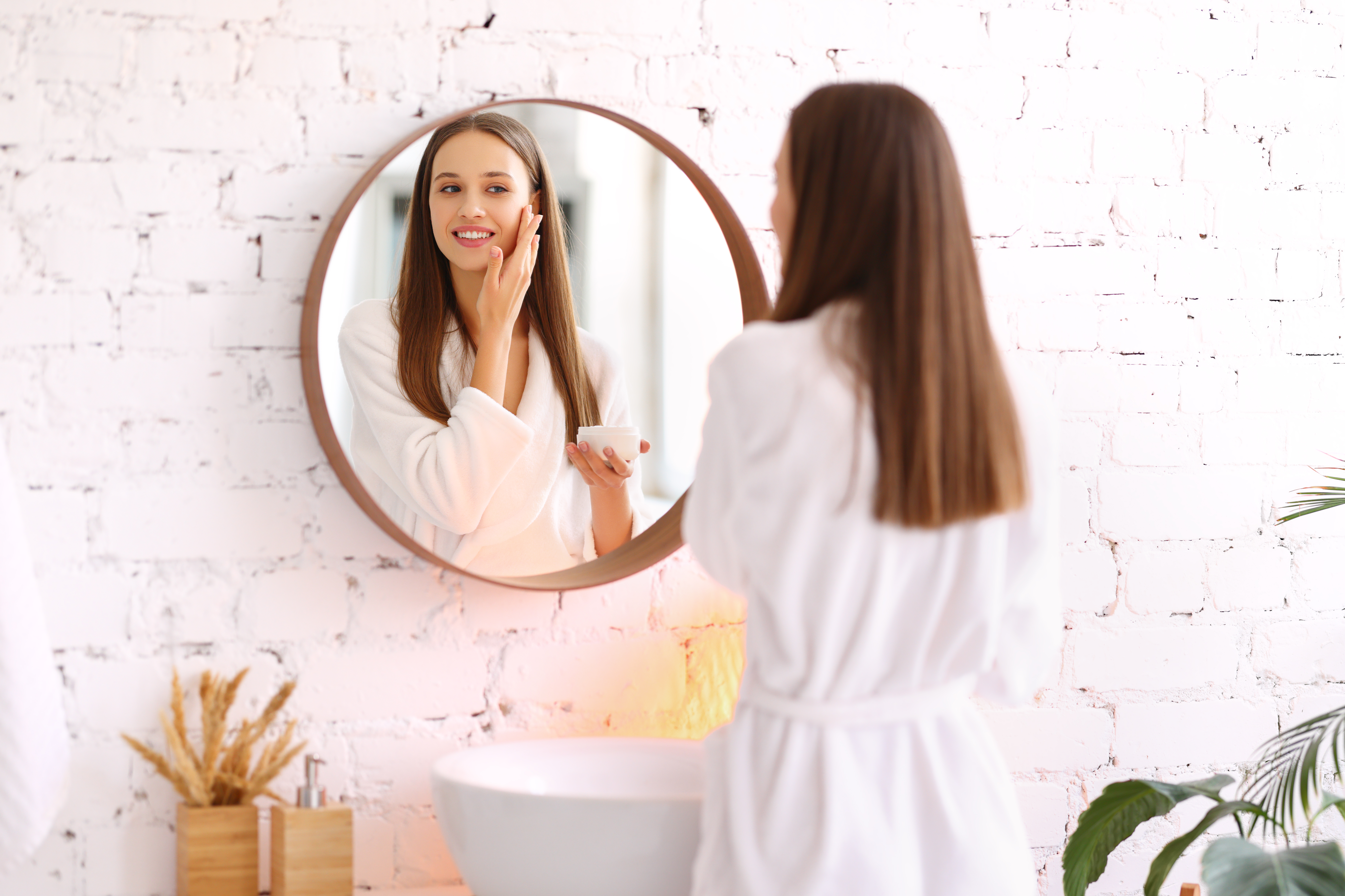 a young woman looks in a mirror while applying balm to her face