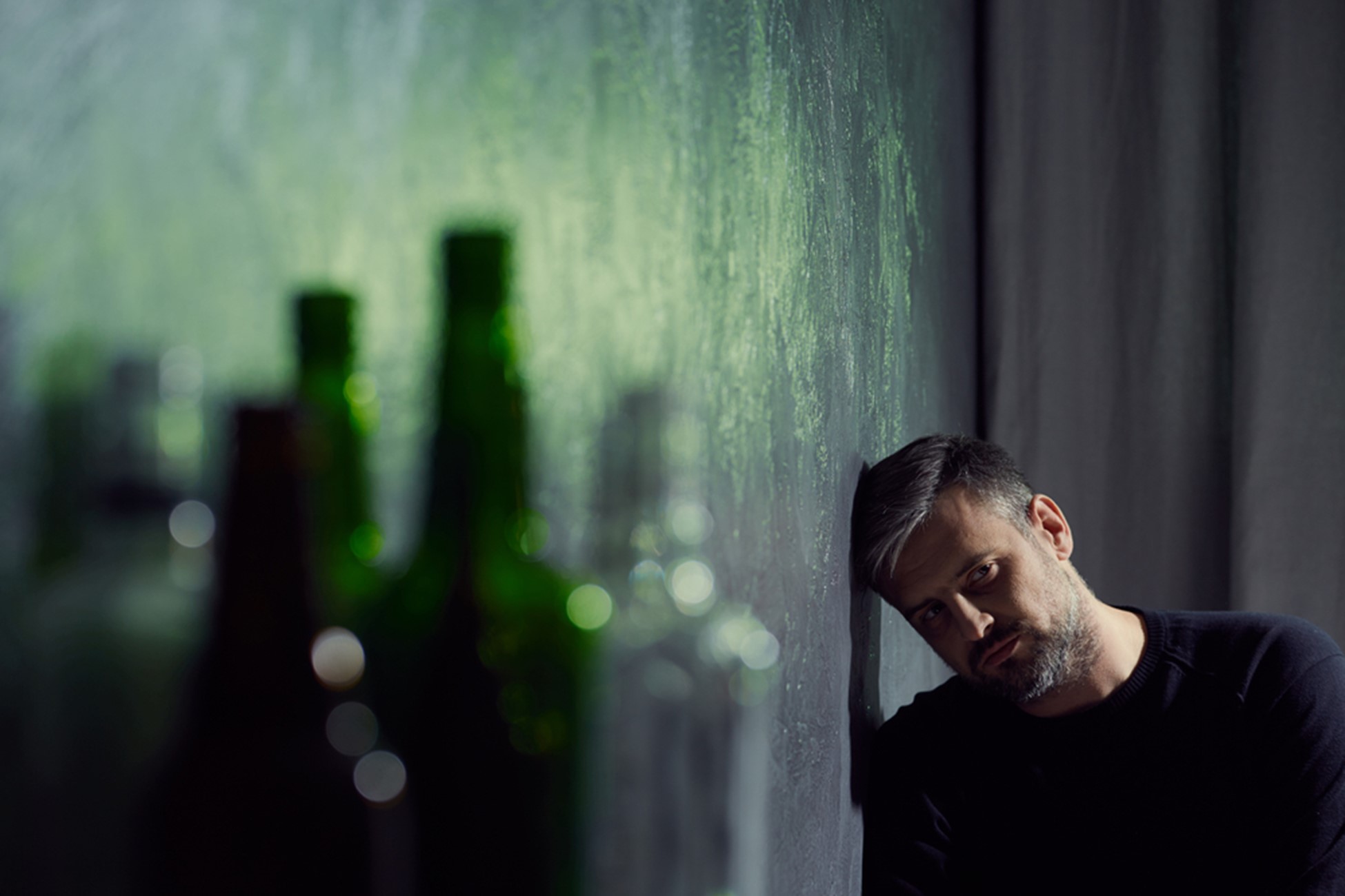 a middle aged man leans with his head against a wall while looking at empty bottles in the foreground