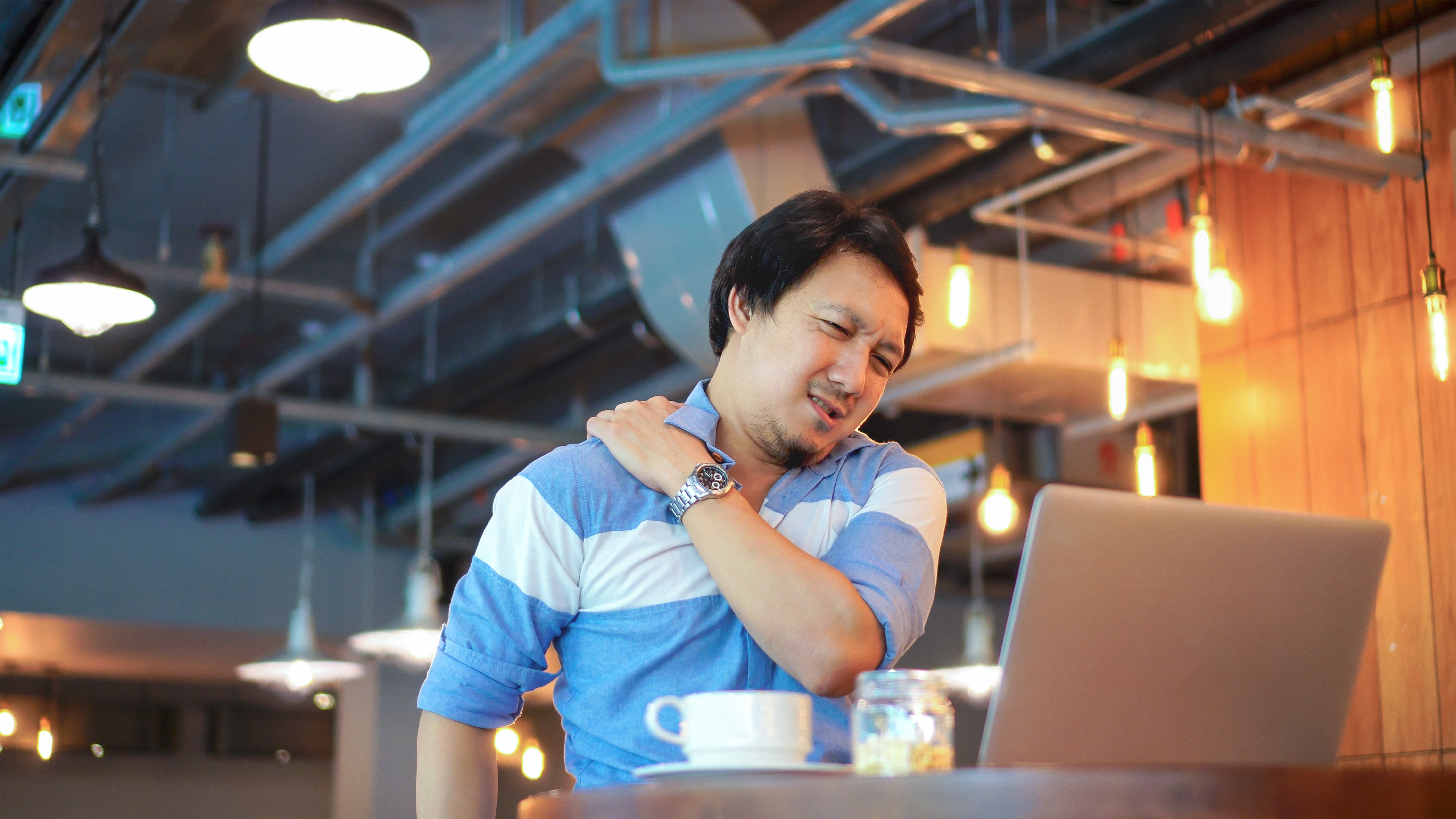 Man working on his laptop grabs at his shoulder and neck in pain