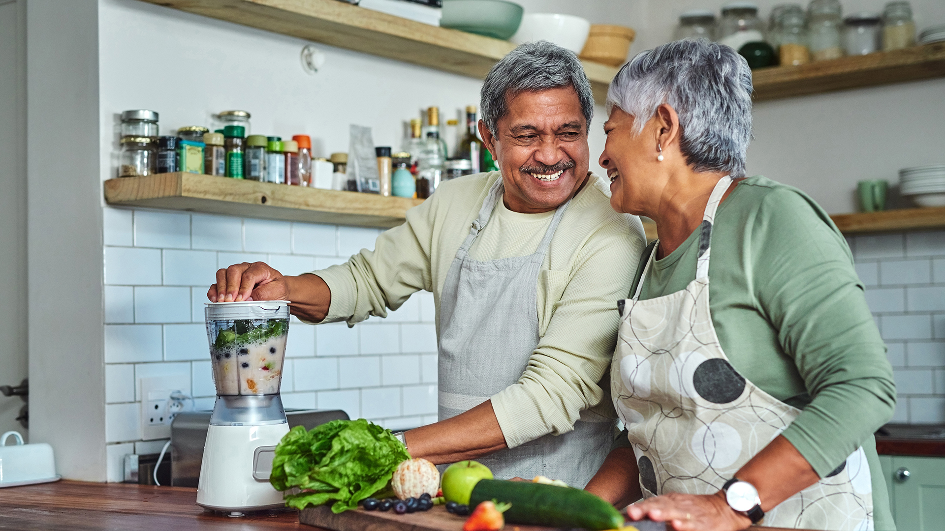 an older couple smiling in the kitchen while cutting vegetables and cooking a healthy meal