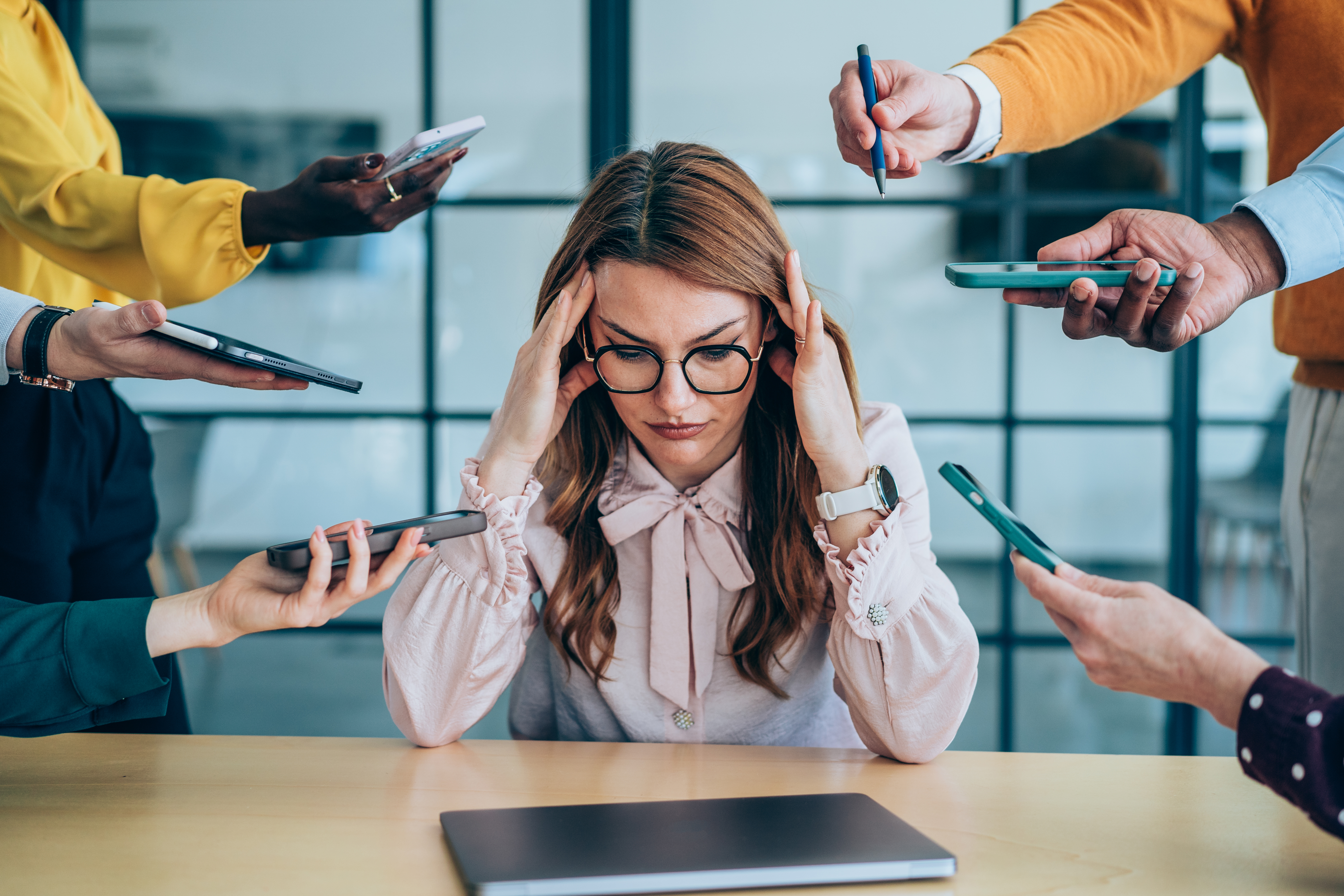 a woman in professional attire is shown massaging her head in distress as several hands are shown reaching toward her with different requests