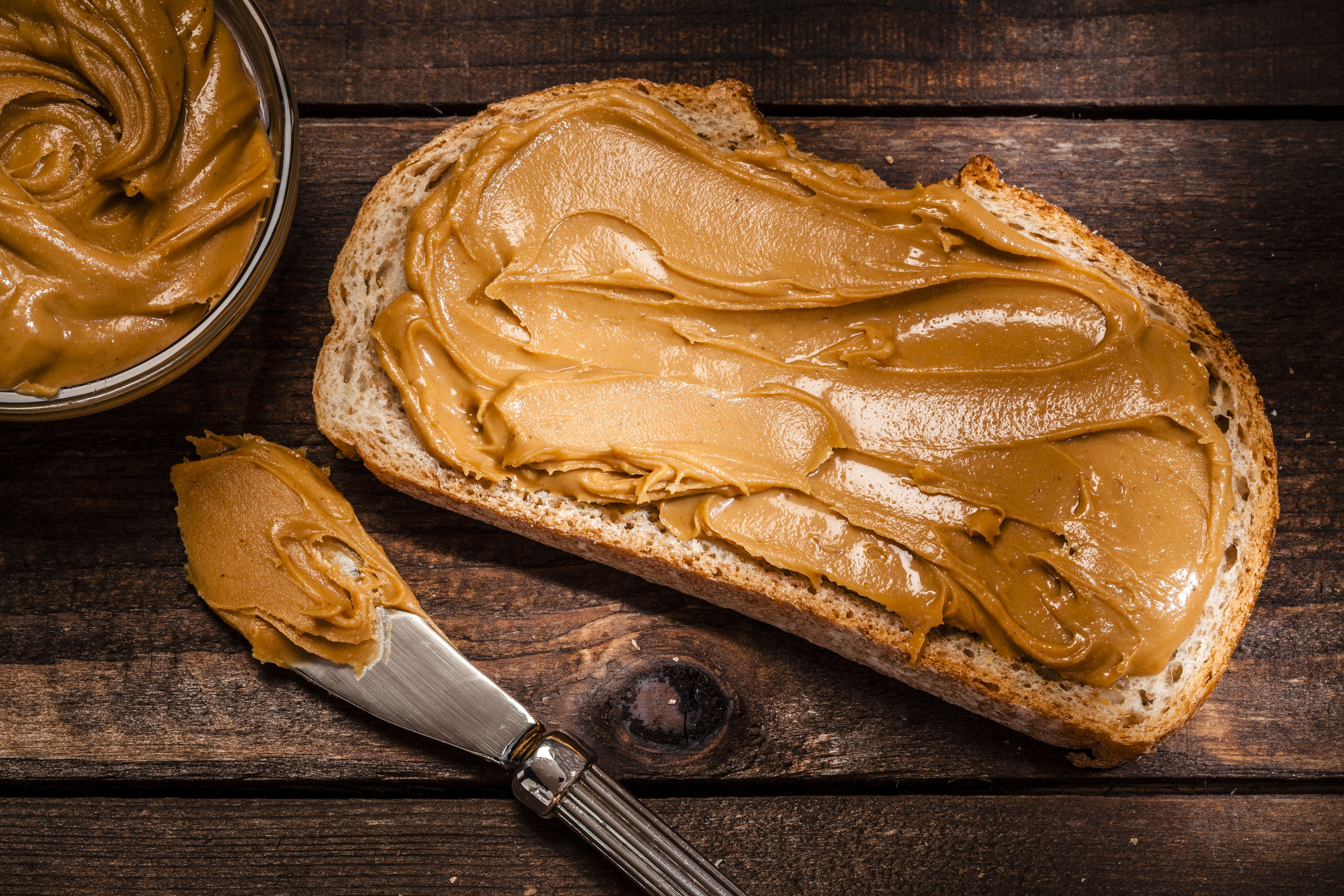 a slice of bread is shown spread with peanut butter, along with the knife used and a jar of peanut butter on top of a wooden table