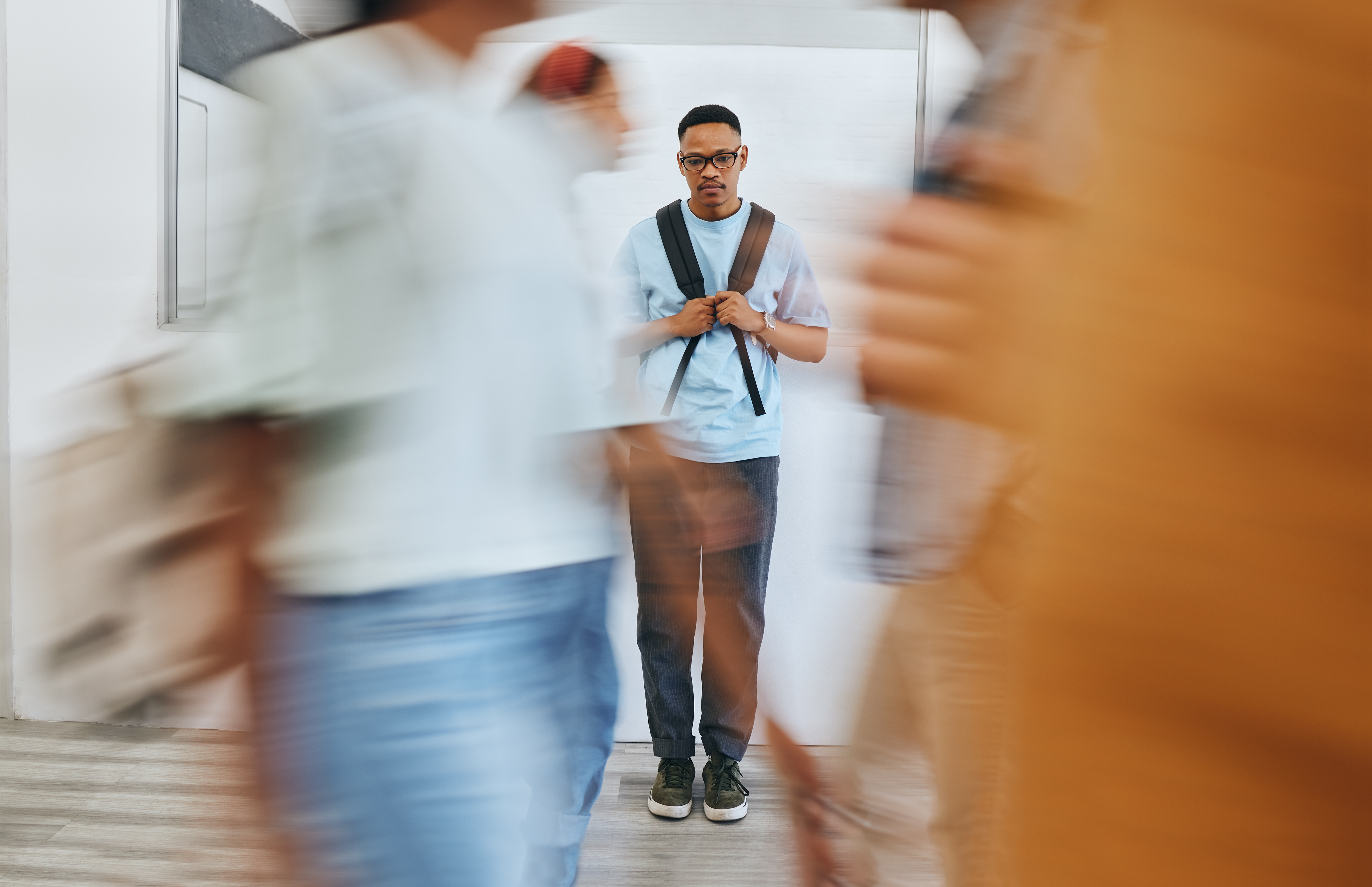 a college aged guy is shown sheepishly wearing a backpack in the background while blurry figures of people walk past in the foreground