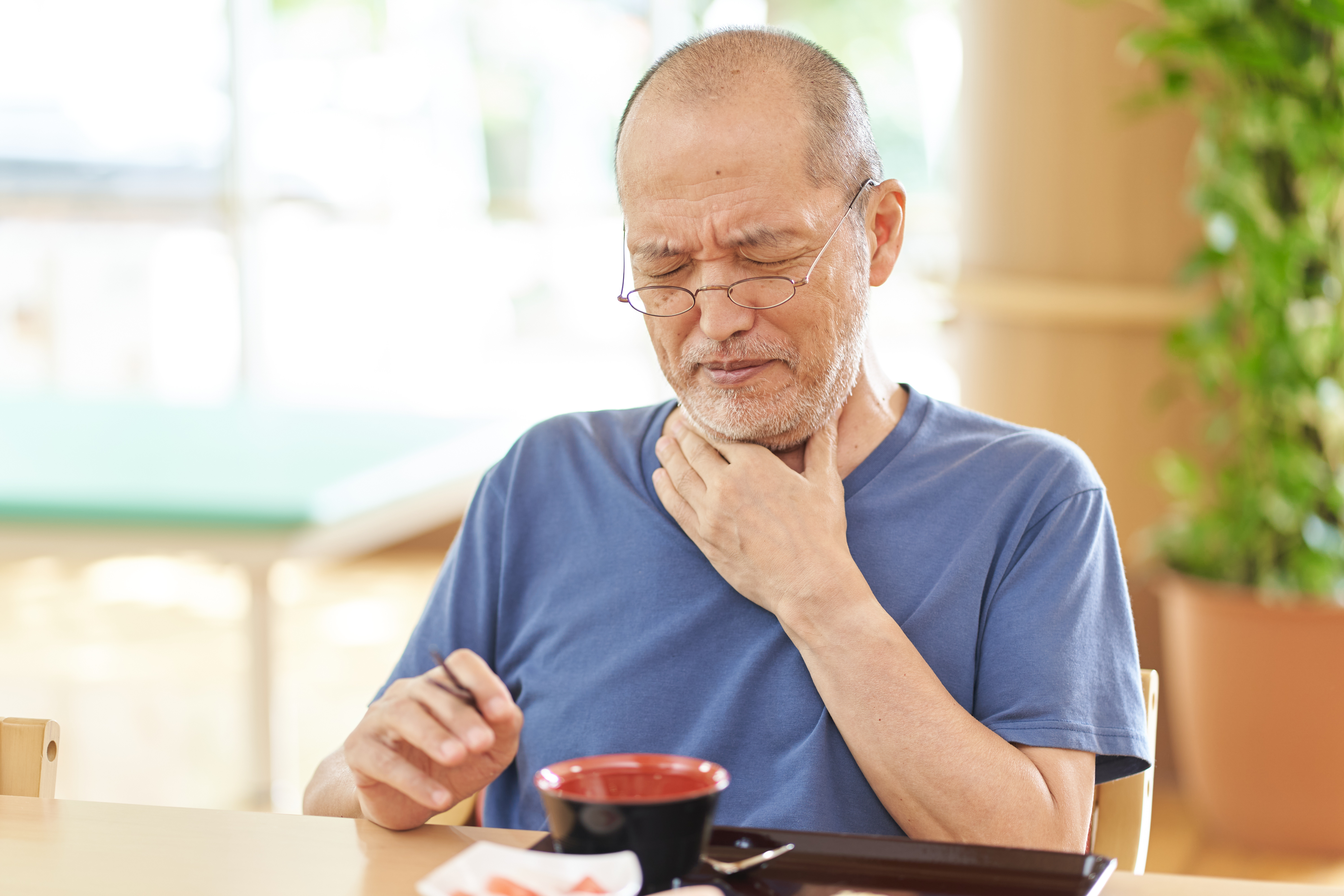an older male is shown sitting at a kitchen table with food and drinks in front of him while he grabs at his upper chest and neck in discomfort