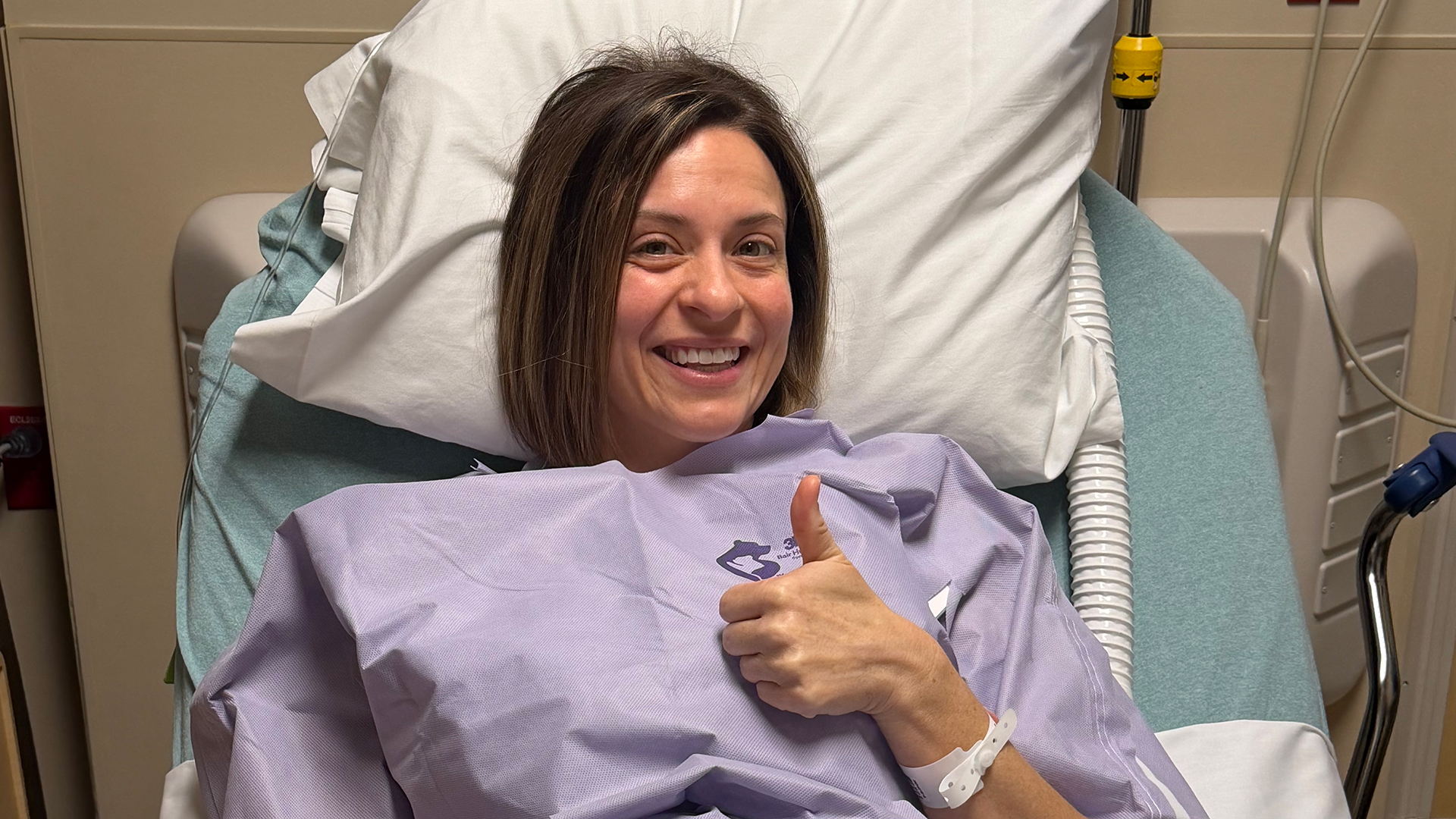 Stefanee is shown giving a thumbs up while in a hospital bed for breast cancer treatment