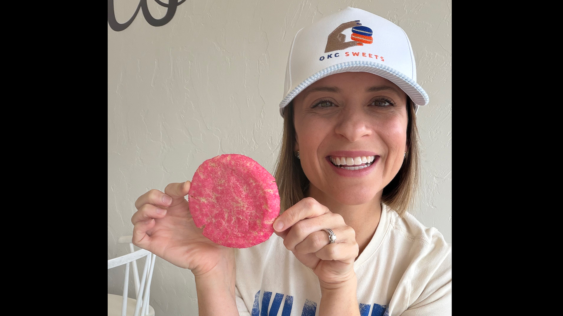Stefanie shown smiling post breast cancer treatment, as she holds up a pink sugar cookie while wearing a white OKC Sweets hat