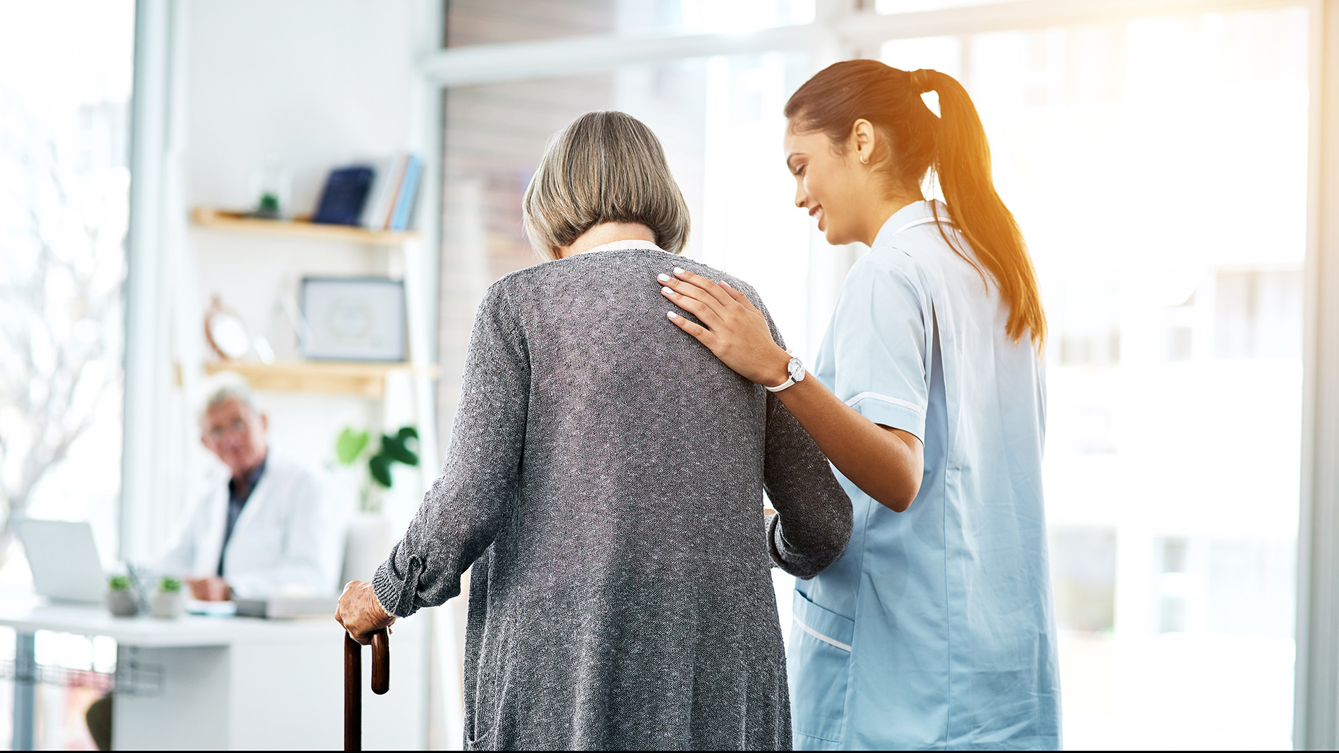 a woman with knee pain uses a cane while being guided by a caregiver and walking into an orthopedic doctor's office 