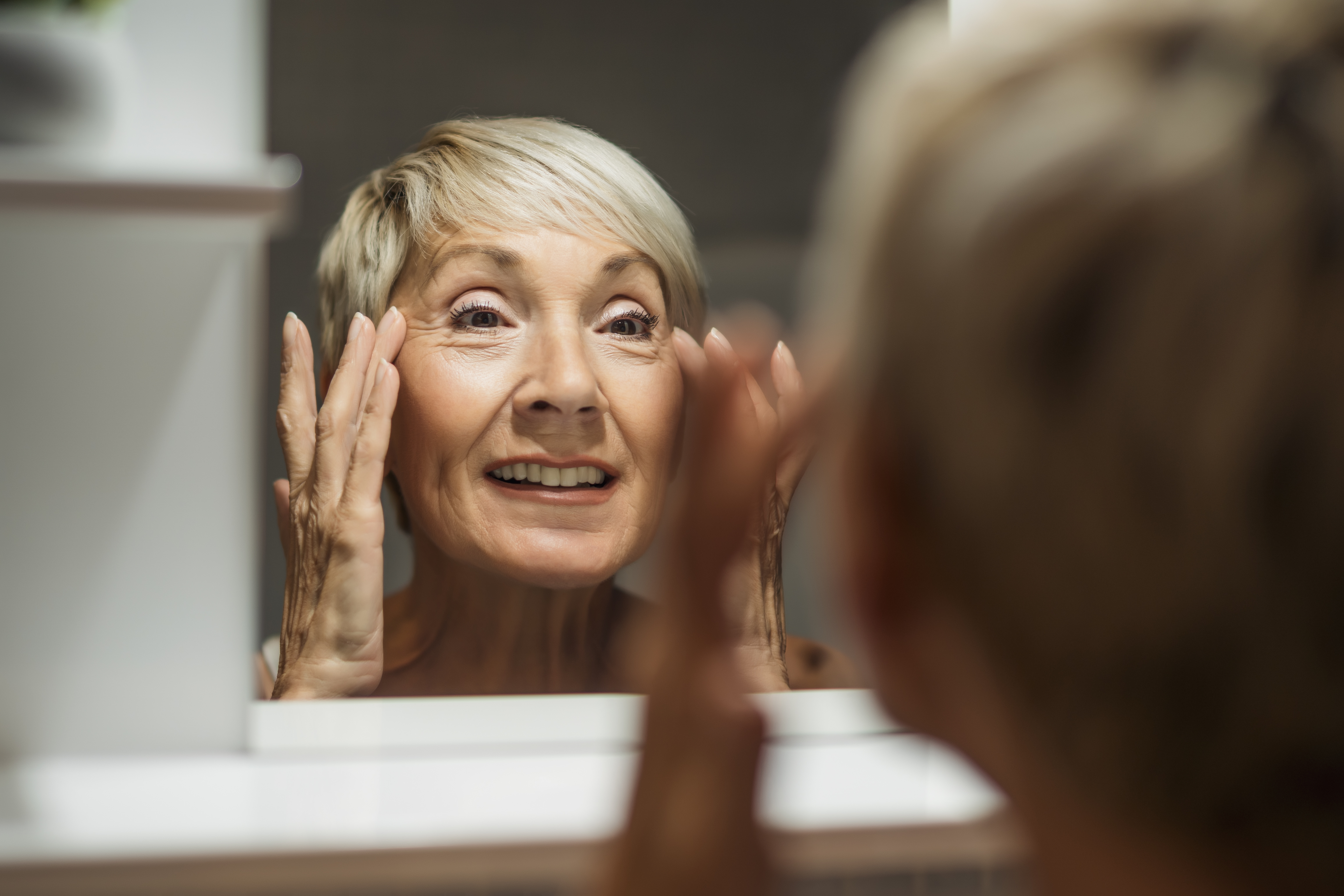 an older woman looks into a mirror with her hands grabbing at the skin around her eyes and face